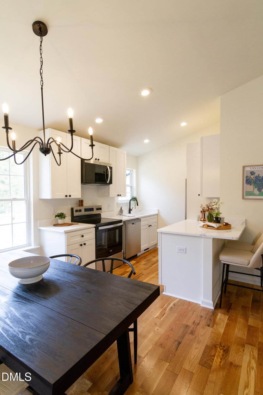 Bright, modern kitchen with white cabinets, stainless steel appliances, and wood flooring. A black dining table with chairs and a bowl sits under a black chandelier. A small breakfast bar with stools is on the right.
