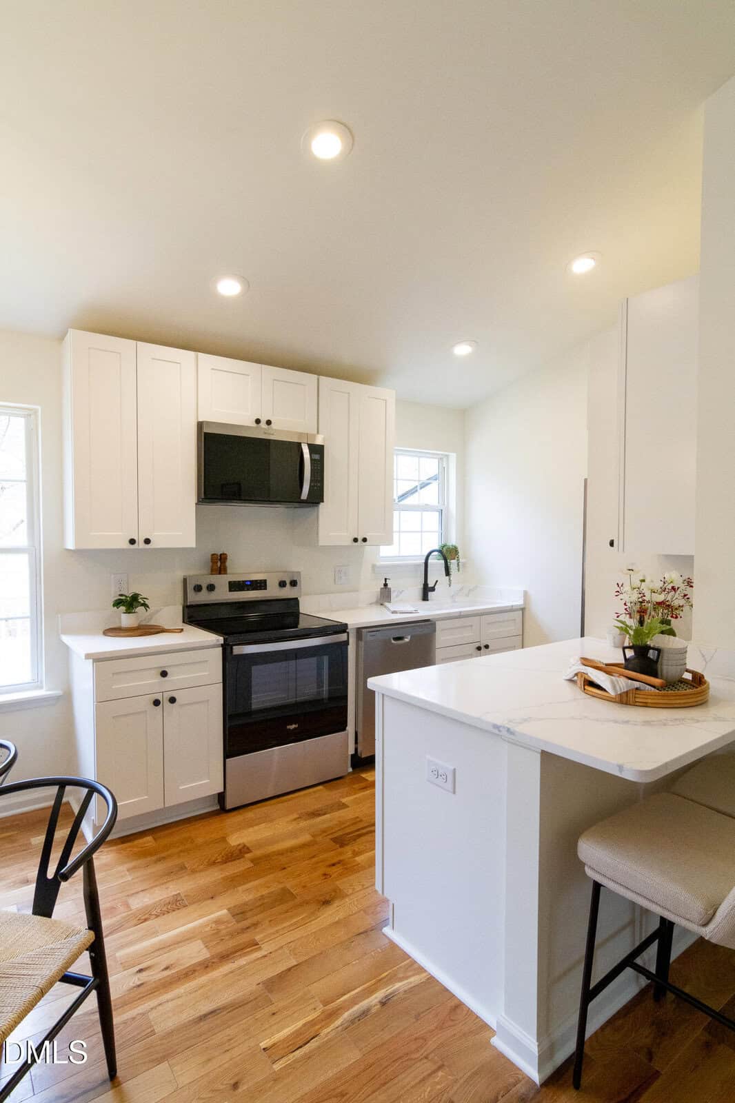 A modern kitchen with white cabinets, a black microwave, black stove, and sink, light countertops, wooden floor, and a small breakfast bar with two beige stools. A plant and decorative tray sit on the counter.