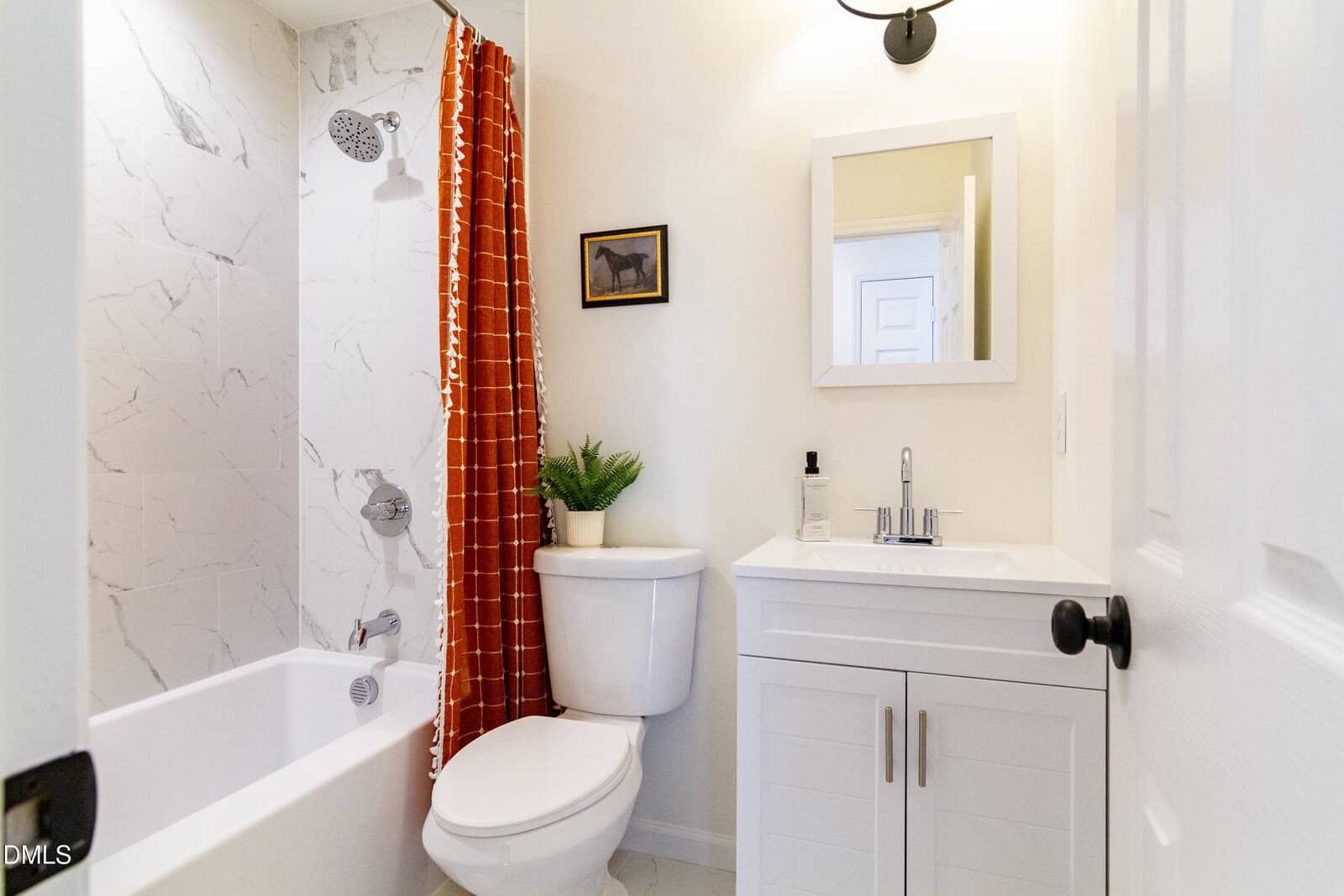 Small bathroom with white walls, a shower-tub with a red curtain, white toilet, white vanity with sink, round mirror, wall light, potted plant, soap dispenser, and a framed horse picture on the wall.