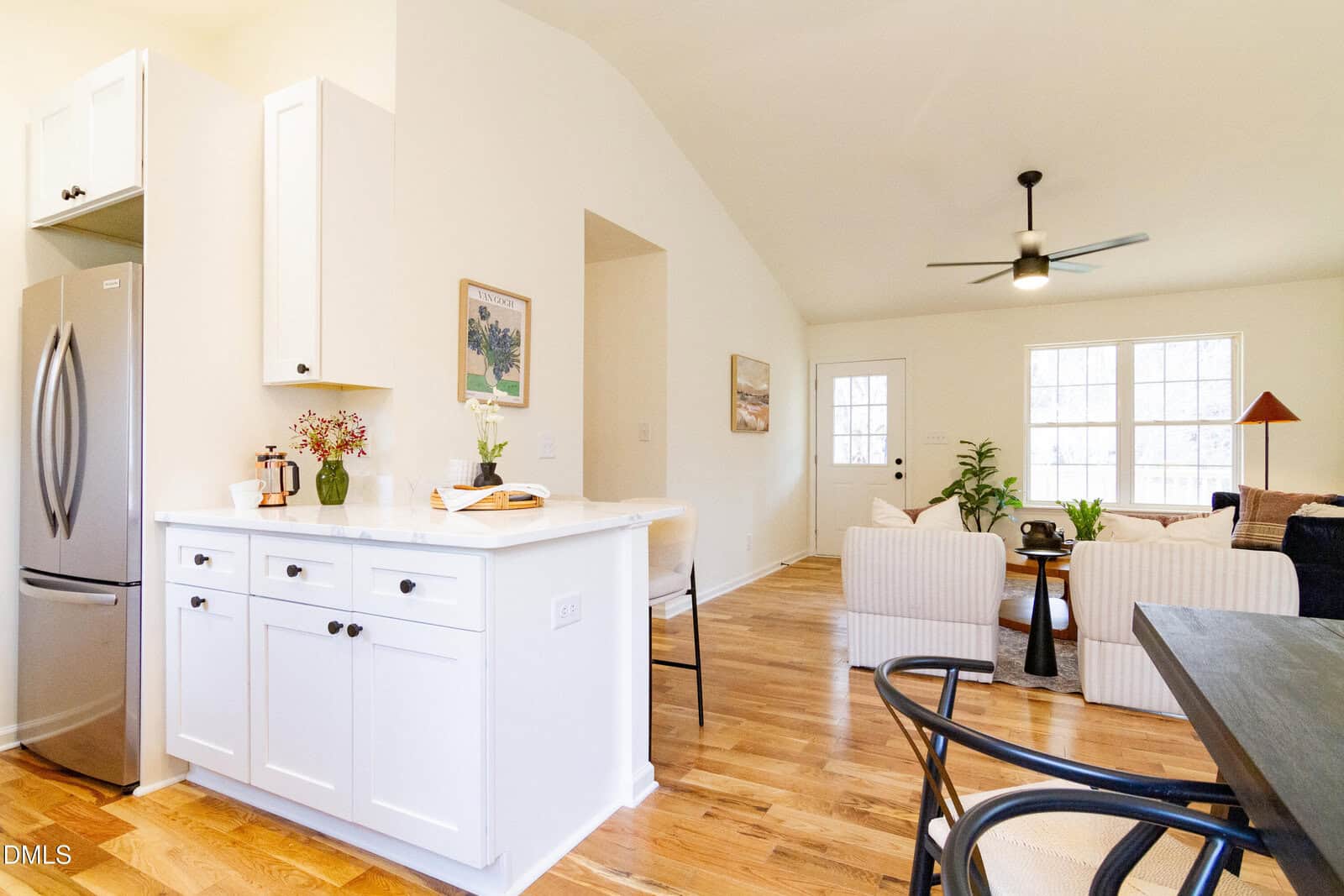 Bright, open-concept living space with white kitchen cabinets, stainless steel fridge, hardwood floors, cozy seating area, large windows, and modern ceiling fan. A plant and artwork add decorative touches.