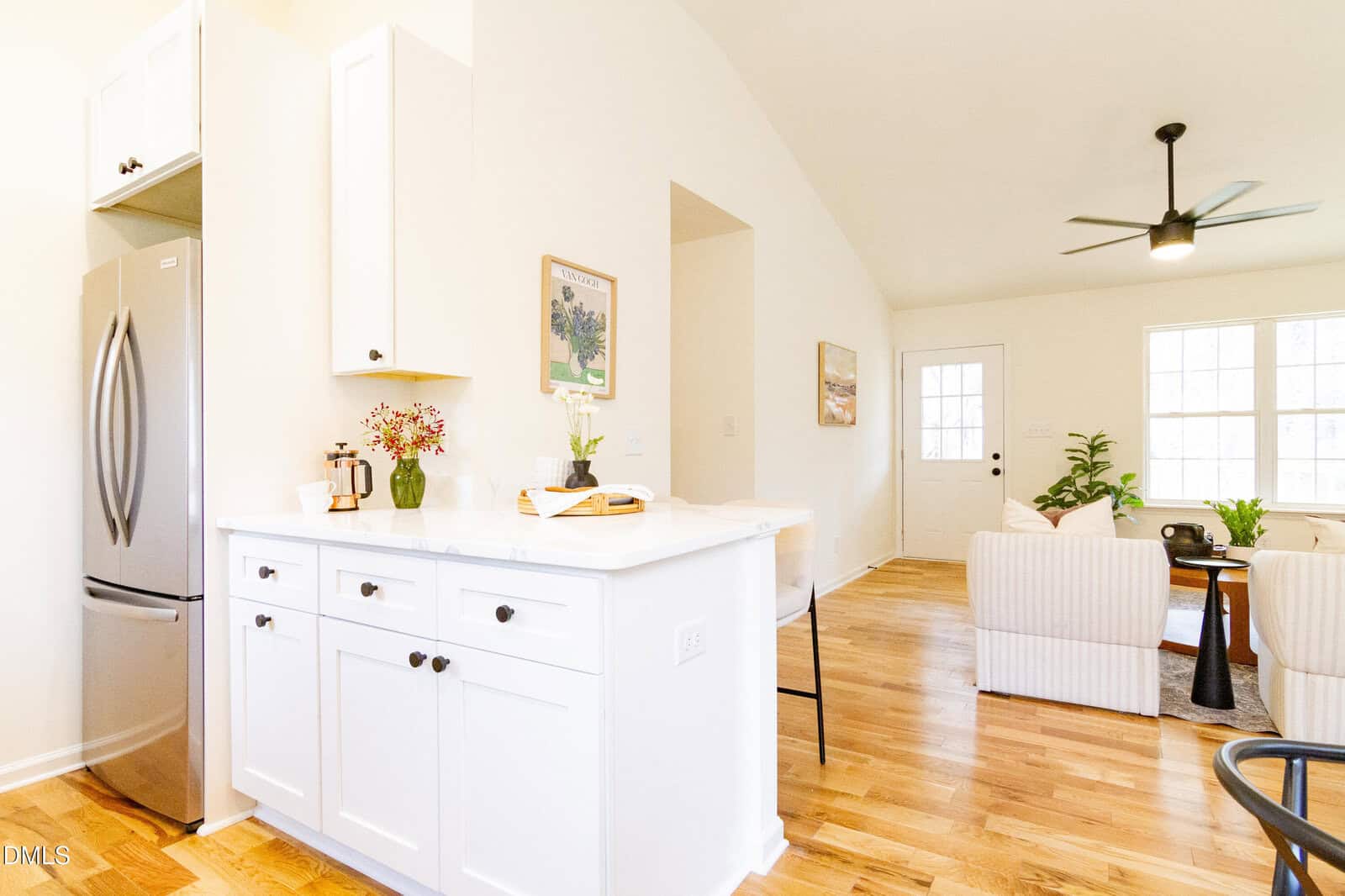 Bright kitchen with white cabinets and a marble countertop, adjacent to a cozy living area with striped armchairs, wooden floors, a ceiling fan, large windows, and a back door letting in natural light.