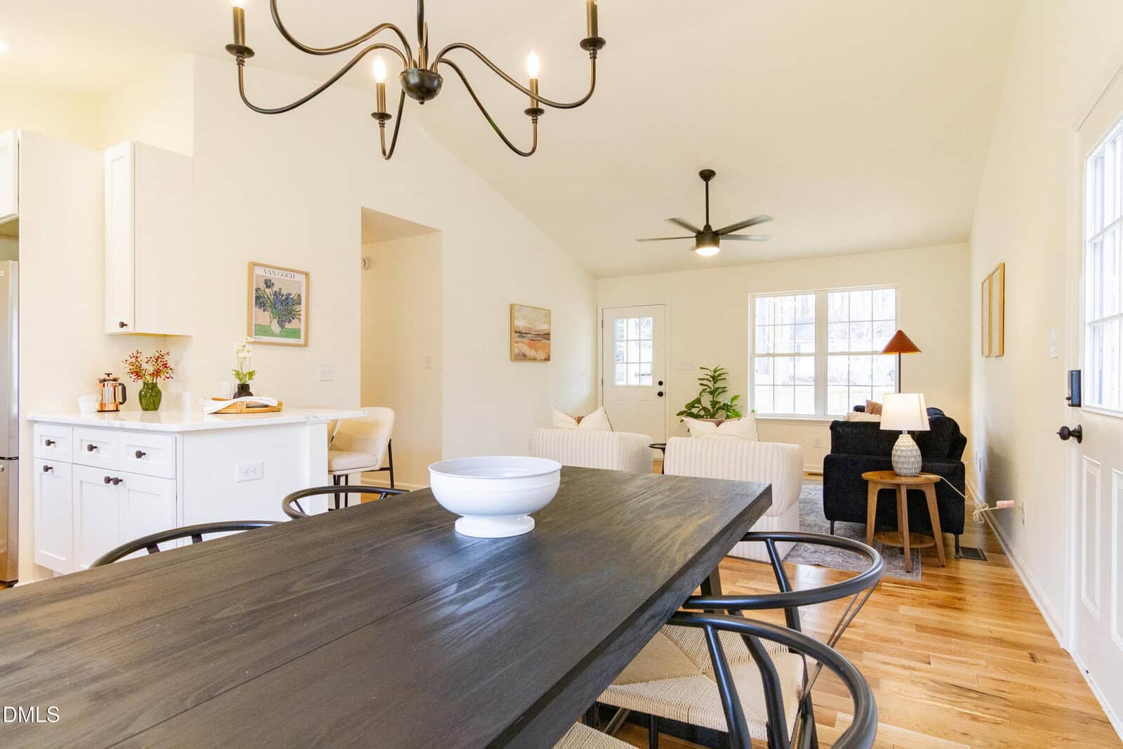 Bright, open-concept dining and living area with white walls, wood floors, black dining table, modern chandelier, and cozy seating by large windows; kitchen with white cabinets and flowers visible on the left.