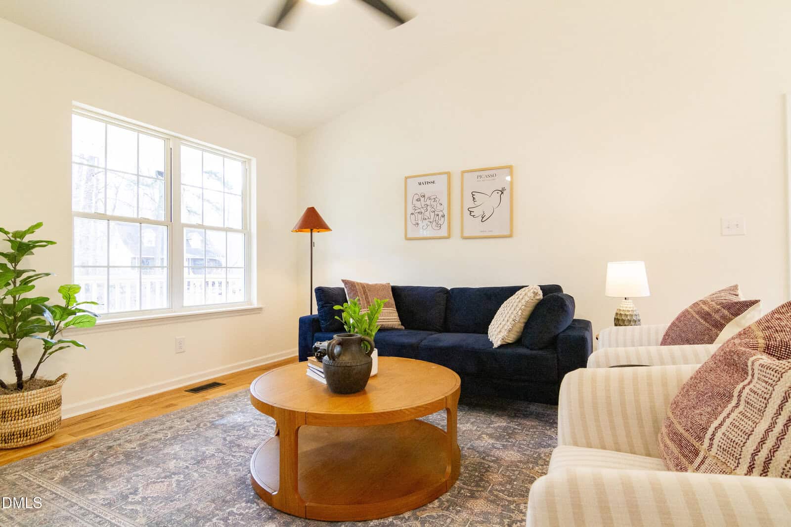 A cozy living room with a navy blue sofa, two striped armchairs, a round wooden coffee table, potted plants, and framed wall art. Large windows let in natural light, brightening the space.