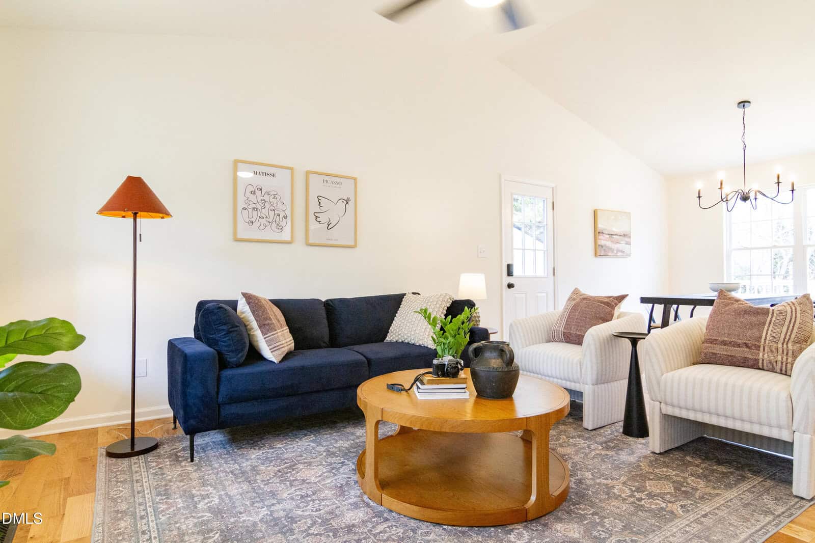 A bright living room with a navy sofa, two white armchairs, a round wooden coffee table, and floor lamp. Art prints hang on the wall, and a dining area with a chandelier is visible in the background.