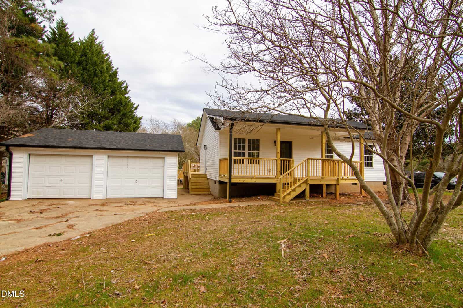 Single-story white house with a covered front porch and wooden steps, next to a detached two-car garage. The yard has some grass and trees, and the sky is overcast.