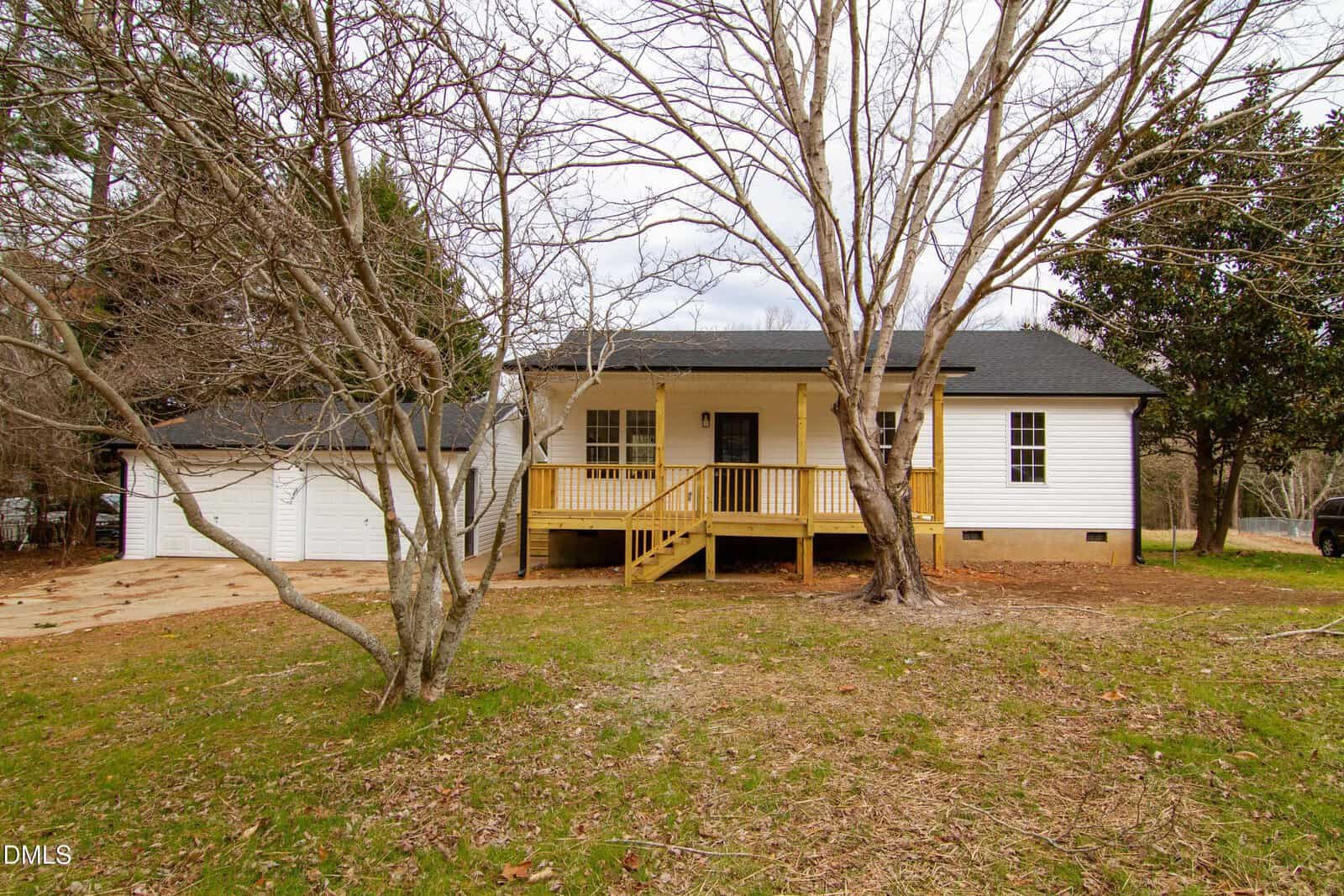 A small white house with black roof, yellow front porch, and attached two-car garage is surrounded by leafless trees and a grassy yard under a cloudy sky.