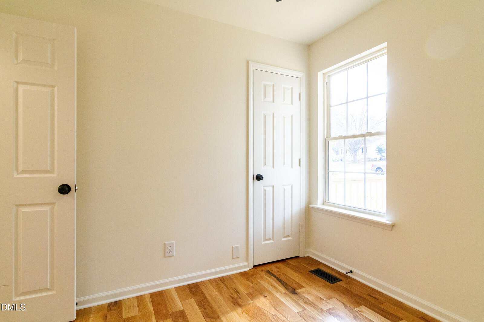 A small, bright room with light beige walls, a hardwood floor, a white door, a closet door, and a large window letting in natural light.