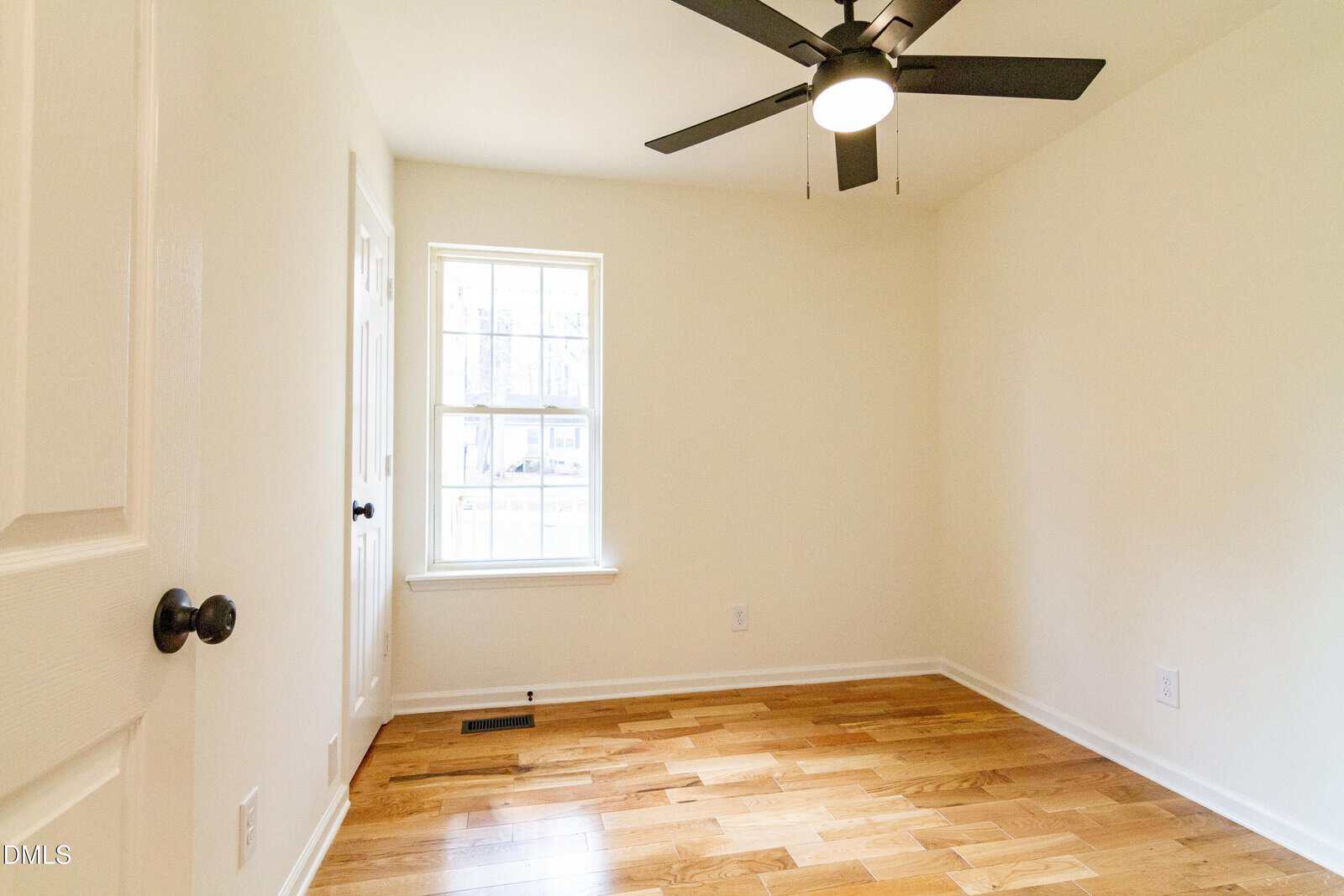 Bright, empty room with light-colored walls, wood flooring, a window letting in daylight, a ceiling fan with a light, and white trim along the baseboards and door.