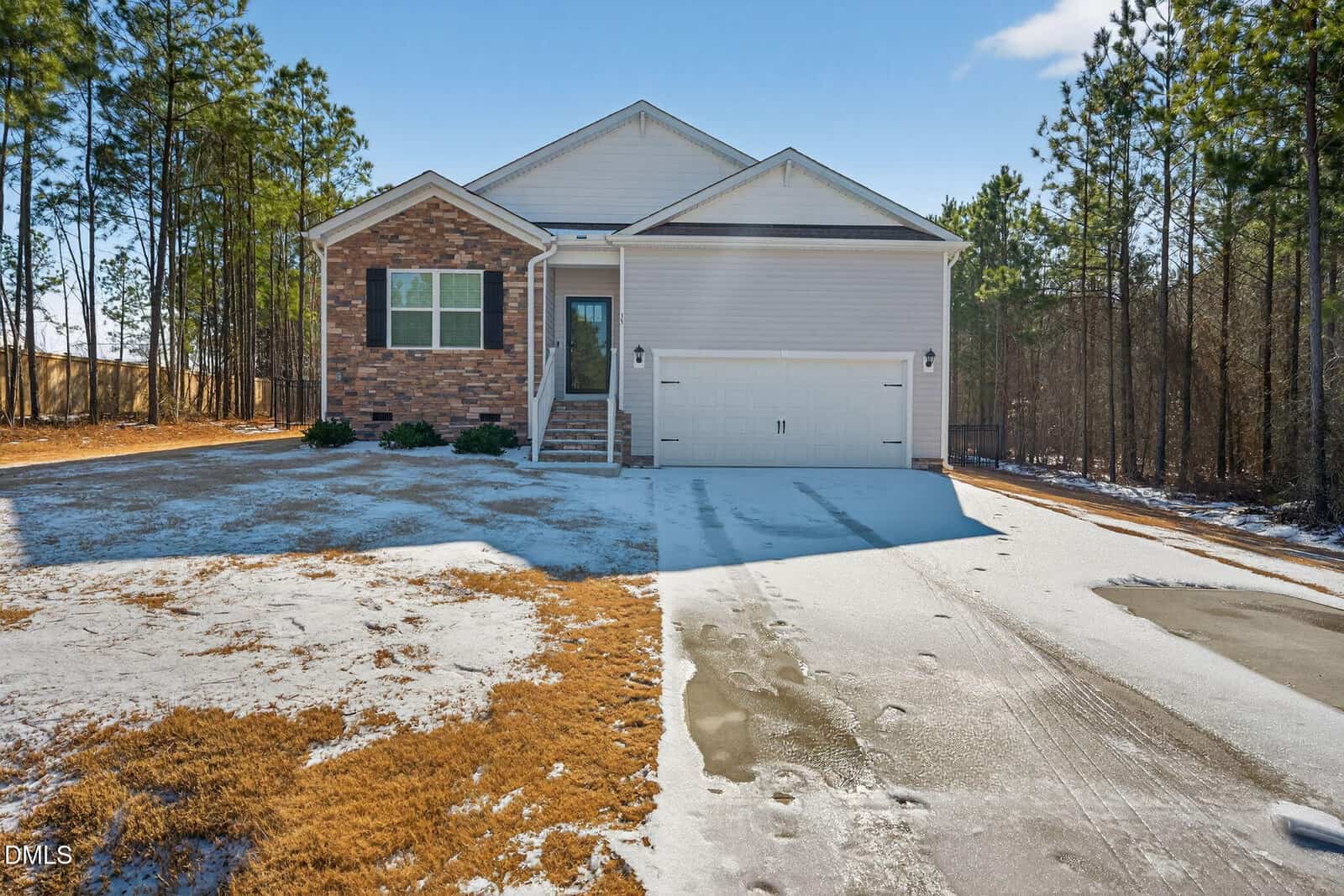 A single-story suburban house with a brick and white siding exterior, a double garage, and a driveway partially covered with snow, surrounded by trees on a sunny day.