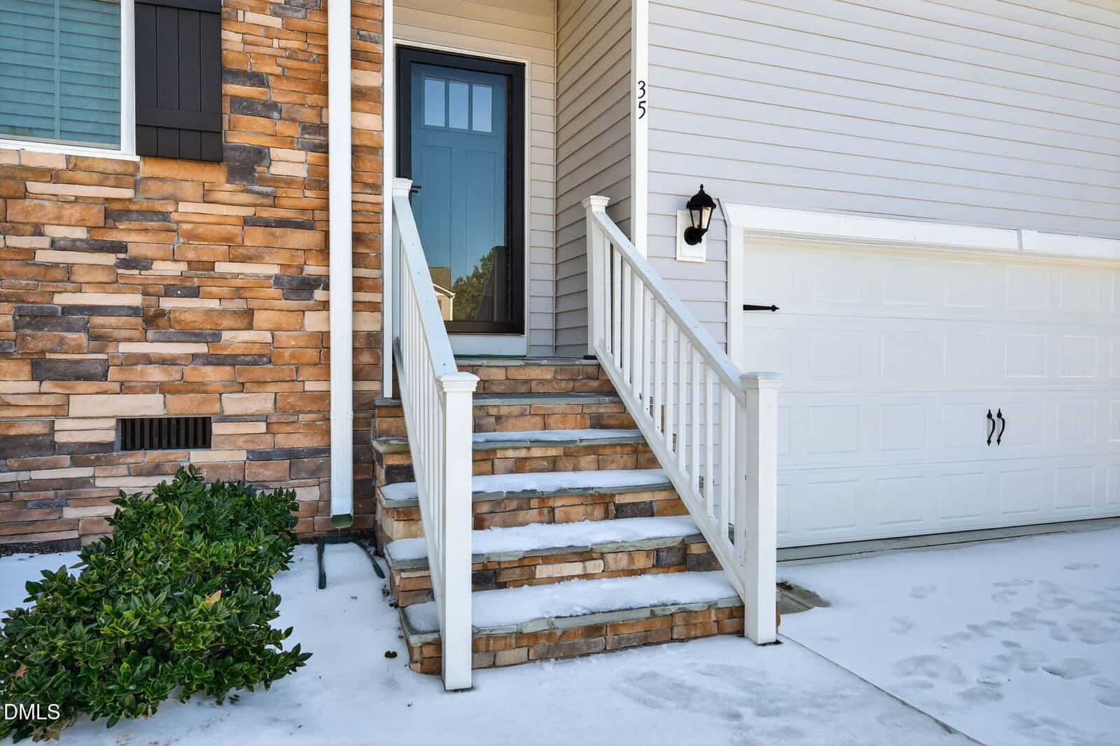 Front steps with a white railing and light snow lead to a black front door of a house with stone and siding exterior, next to a white garage door. A small bush grows beside the steps.