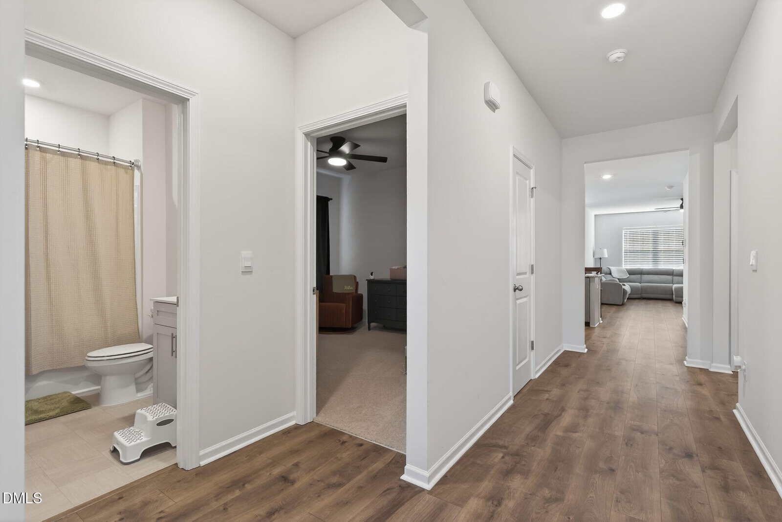 A bright hallway with wood floors leads to a bathroom with a beige shower curtain, a bedroom with carpet and a ceiling fan, and a living room with a sofa and large windows.