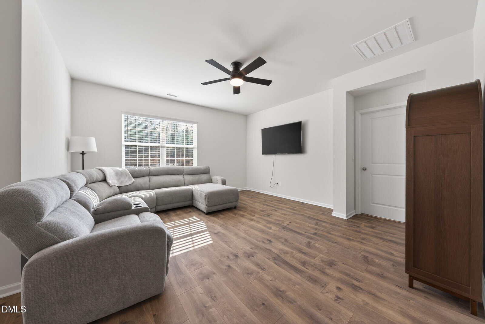 A bright living room with a gray sectional sofa, a floor lamp, wall-mounted TV, wood flooring, and white walls. Sunlight streams in through a large window with blinds.