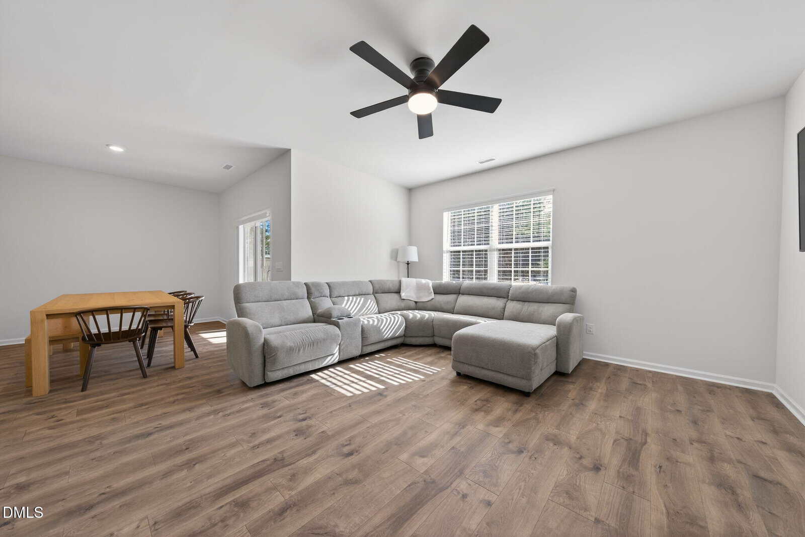 A bright living room with wood flooring, a gray sectional sofa, a ceiling fan, large window with blinds, and a wooden dining table with chairs in the adjacent dining area.