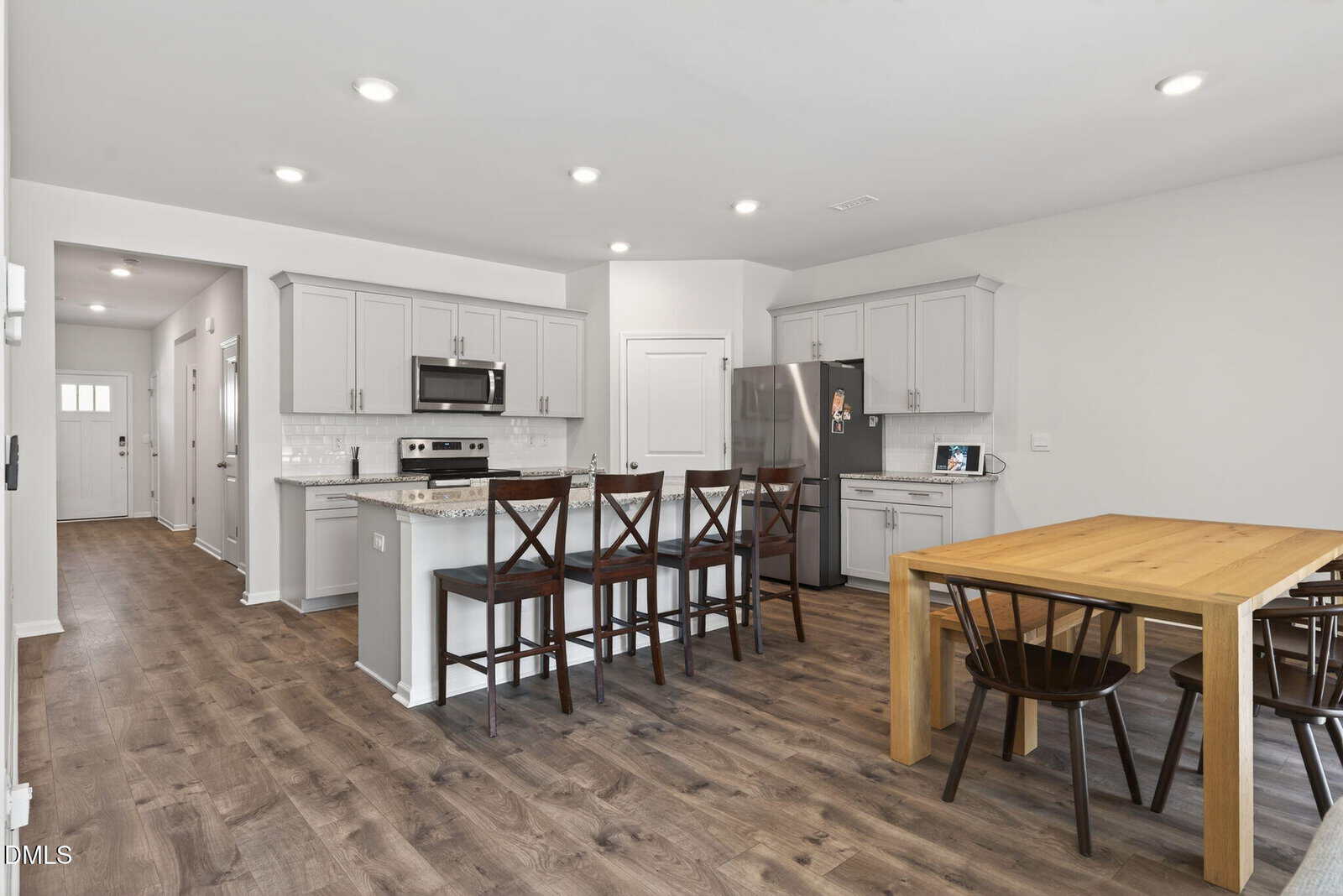 Modern kitchen and dining area with wood flooring, light gray cabinets, stainless steel appliances, a kitchen island with four barstools, and a wooden dining table with four chairs. Bright, open, and minimally decorated.
