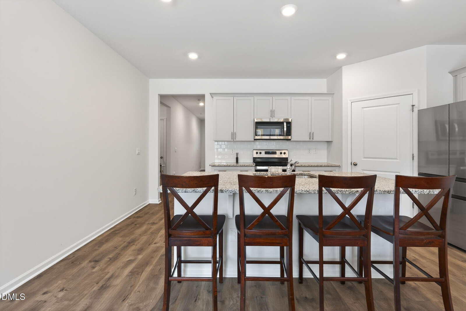 A modern kitchen with white cabinets, stainless steel appliances, granite countertops, and four dark wooden bar stools at a breakfast bar on wood flooring.