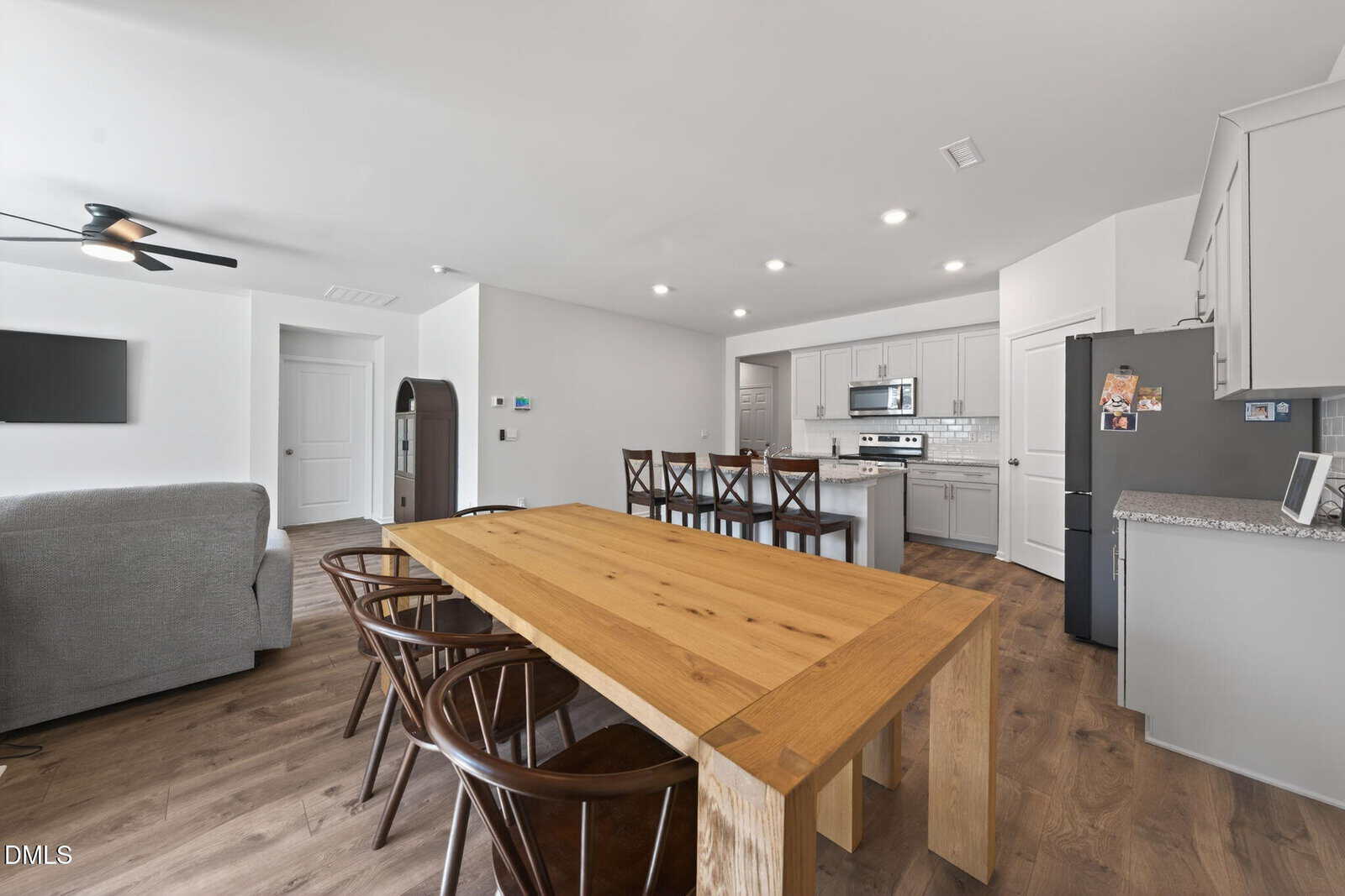 Modern open-concept kitchen and dining area with a wooden dining table, six chairs, kitchen island with barstools, gray cabinets, stainless steel appliances, and a cozy living space with a sofa and wall-mounted TV.