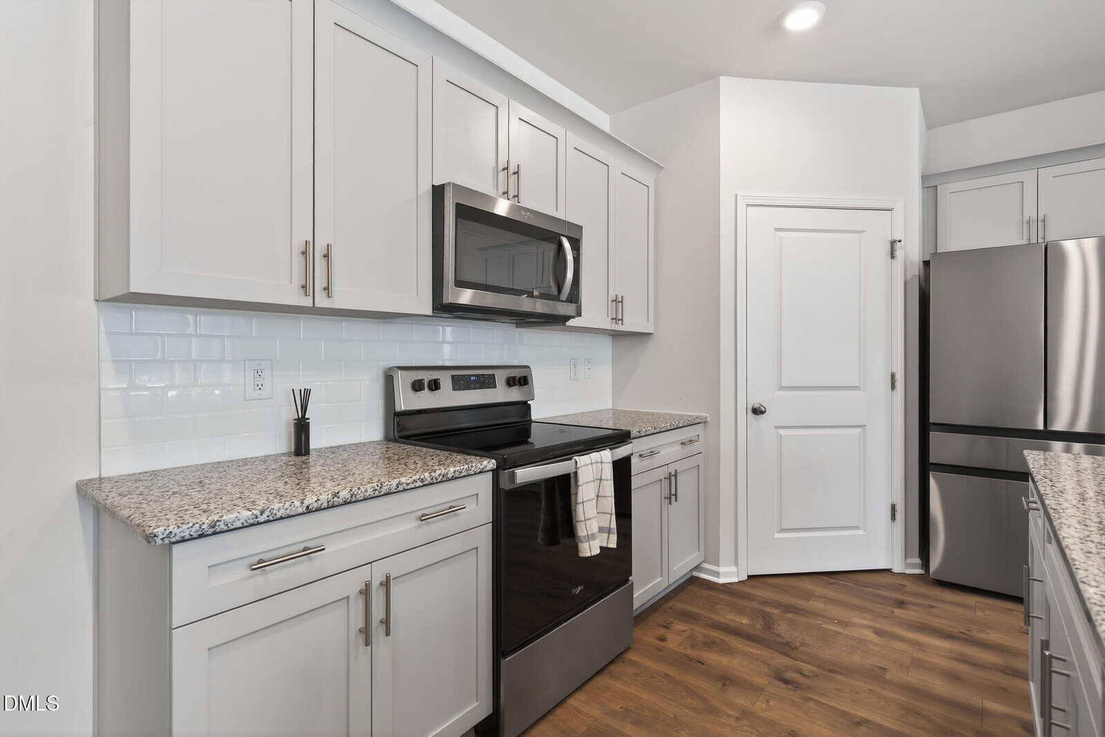 Modern kitchen with white cabinets, stainless steel appliances, granite countertops, subway tile backsplash, electric stove, and a microwave. Wooden floor and a white door are also visible. A striped towel hangs on the oven handle.