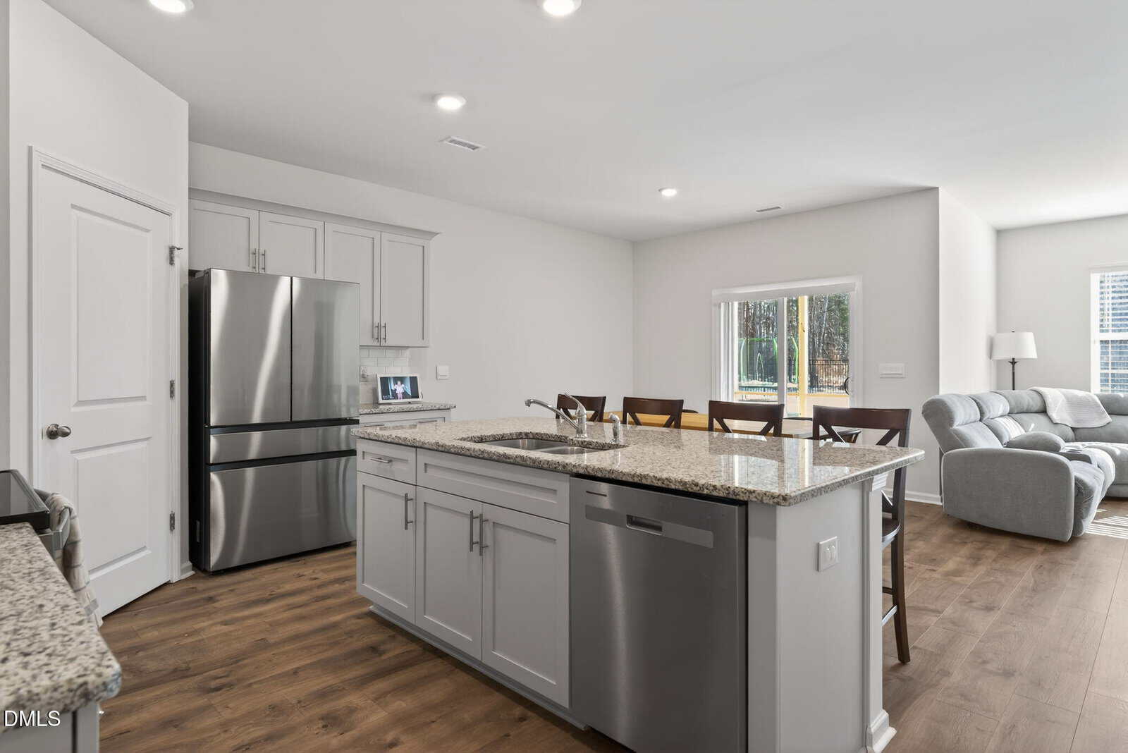 Modern kitchen with granite countertops, stainless steel appliances, and an island with a sink and dishwasher. Four wooden chairs line the island, and a gray sofa is visible in the adjacent living area.