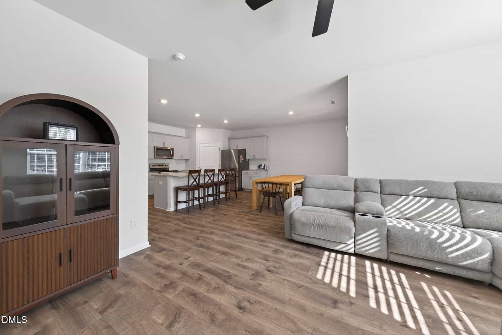 Open-plan living area with wood floors, a gray sectional sofa, wooden dining table with chairs, kitchen with island seating, white cabinets, and a dark wooden cabinet on the left. Sunlight streams in through window blinds.