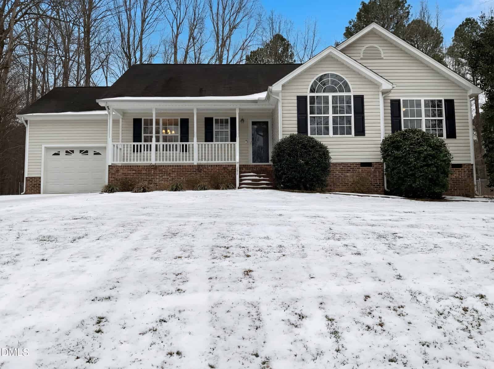 A light yellow house with black shutters, a front porch, and attached garage sits on a snow-covered lawn with bare trees in the background. Large bushes flank the front steps leading to the porch.