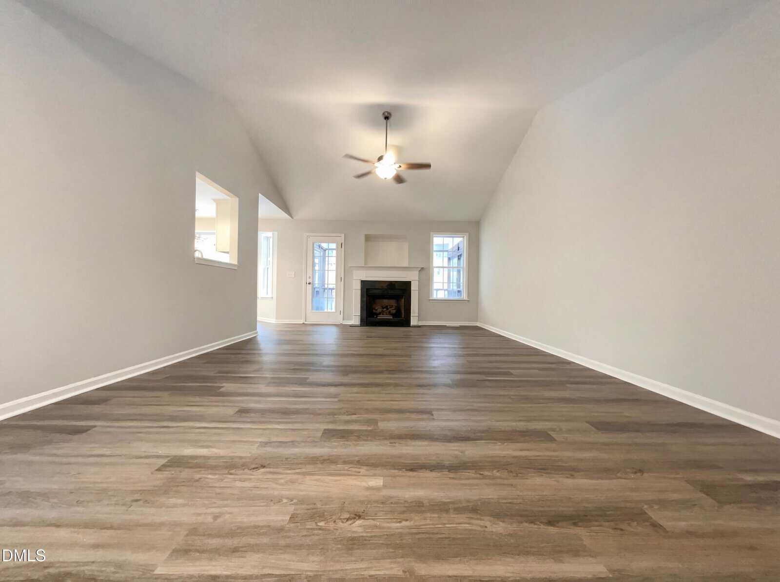 A spacious, empty living room with light gray walls, wood-style flooring, a ceiling fan with light, a fireplace, and two large windows on either side of the fireplace. An open doorway leads to another room.