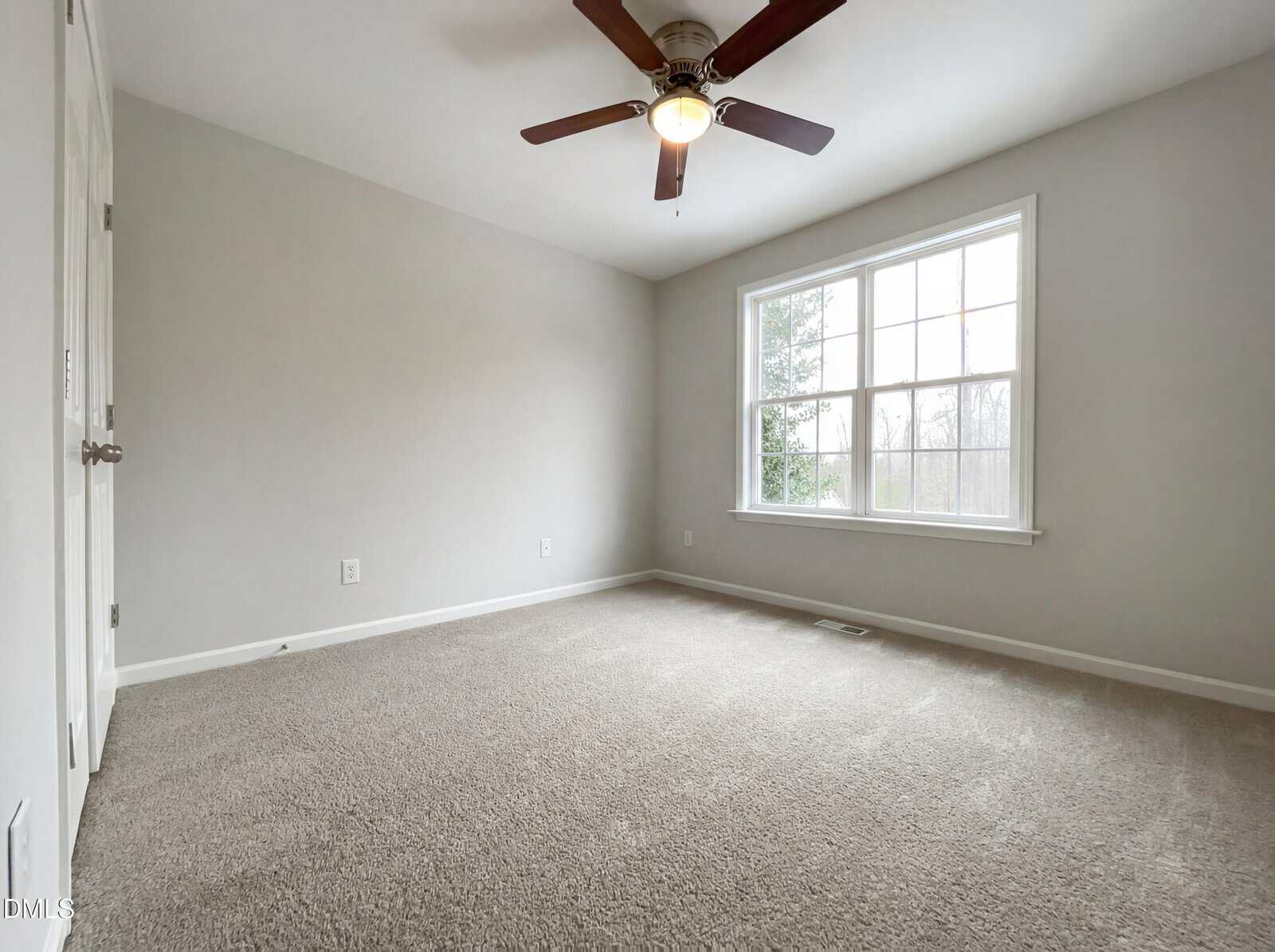 A bright, empty room with light gray walls, beige carpet, a ceiling fan with a light, and a large window letting in natural light.
