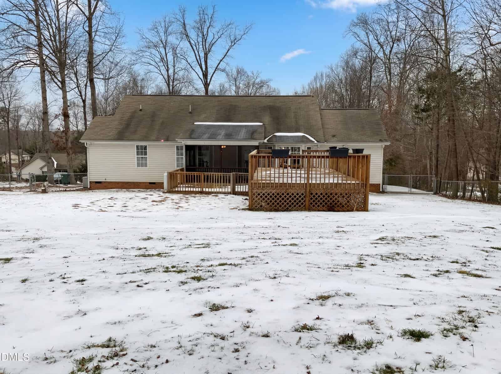 A one-story house with a wooden deck sits in a fenced backyard lightly covered with snow. Leafless trees surround the yard under a partly cloudy blue sky.