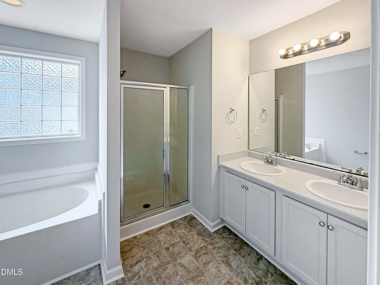 A modern bathroom with a glass shower, a white bathtub beneath a glass block window, double sinks with a large mirror, and light fixtures above. The floor is tiled and cabinets are white.