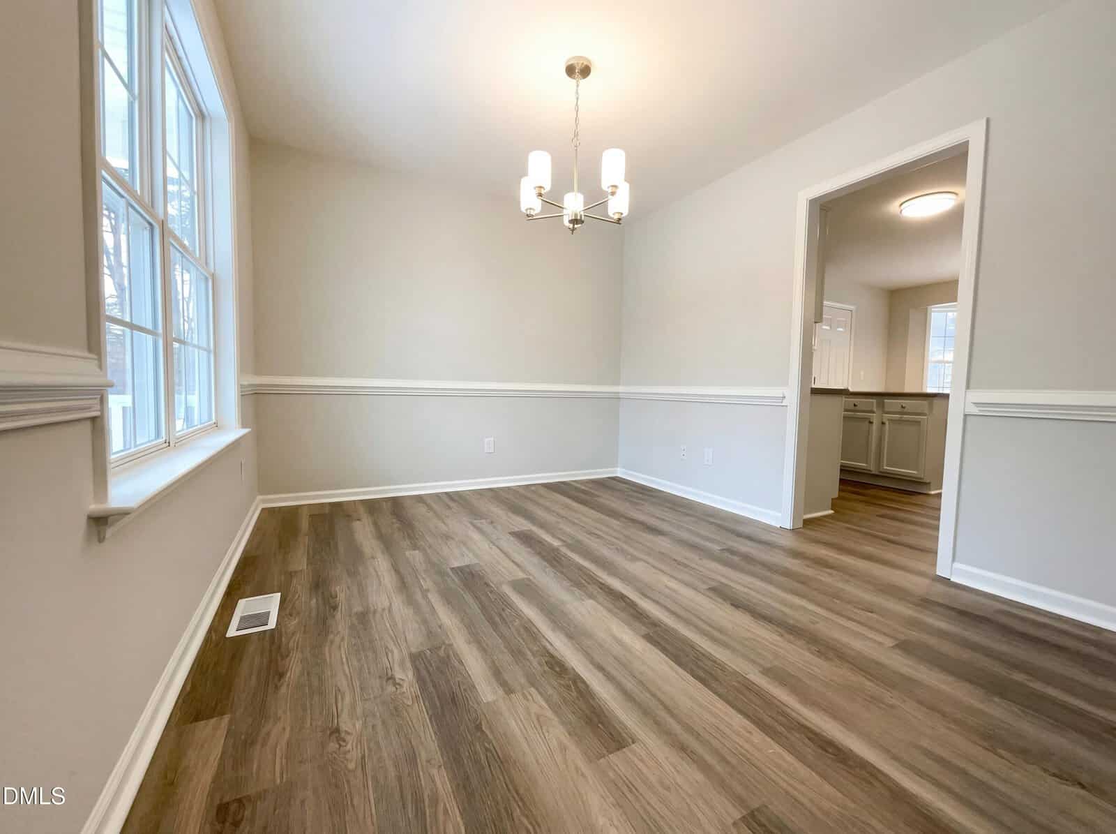 A bright, empty dining room with light gray walls, wood-look flooring, large window, and modern chandelier. An open doorway leads to a white kitchen in the background.