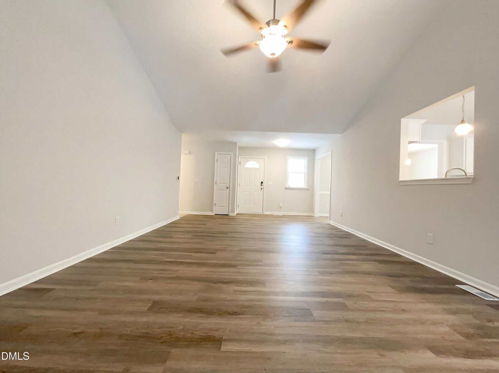 Spacious, empty living room with light gray walls, wood-style flooring, ceiling fan with light, and a windowed opening to the kitchen on the right. White doors and trim are visible at the far end.