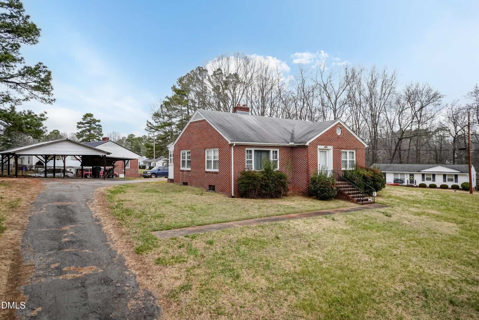 A small red brick house with white trim sits on a grassy lot with a paved driveway. There are steps leading to the front door and a carport with multiple vehicles in the background. Trees and another house are visible nearby.