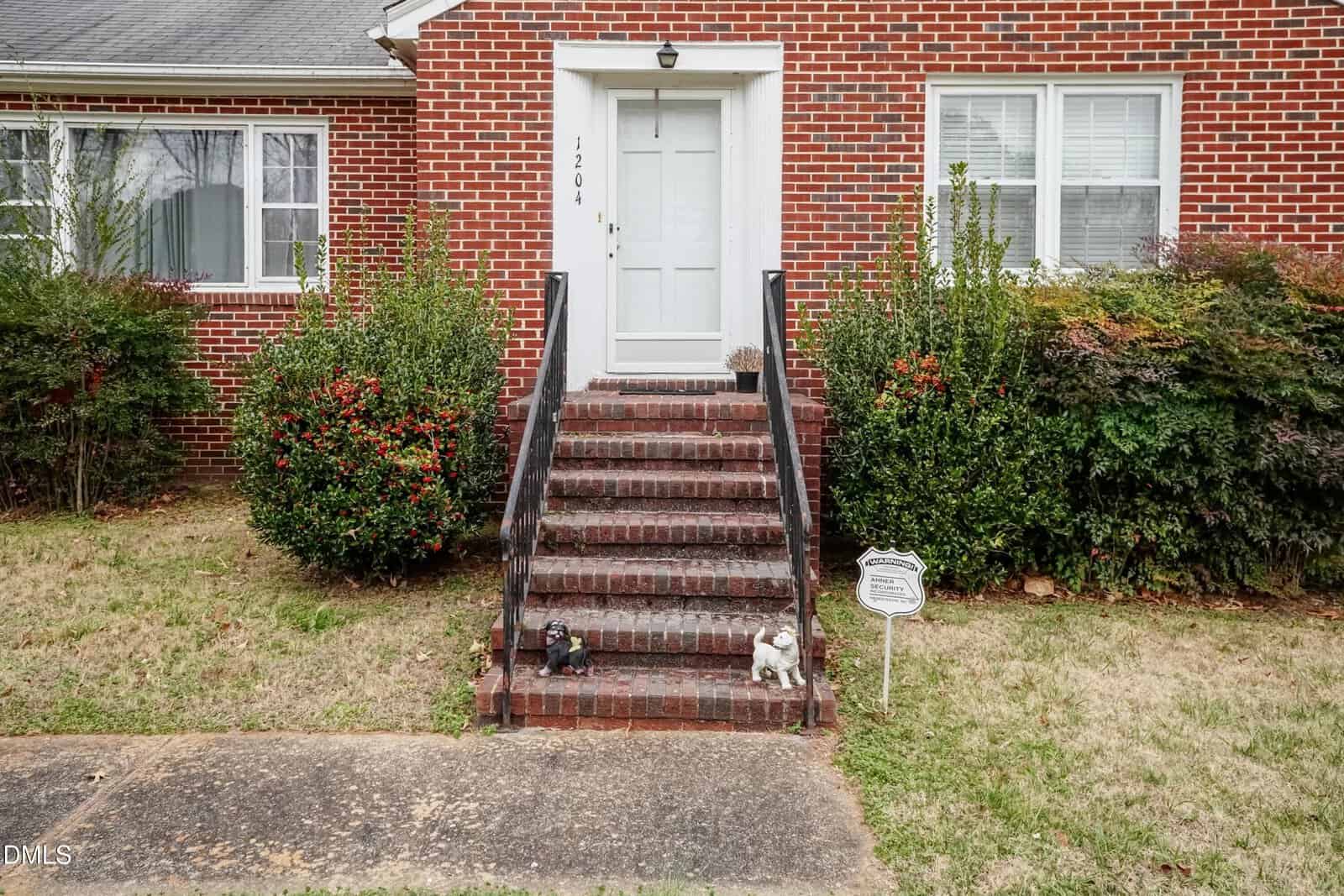 Brick steps with black railings lead to a white front door of a red brick house. Bushes flank the steps, and two small animal statues and a white sign are on the bottom step near the sidewalk.