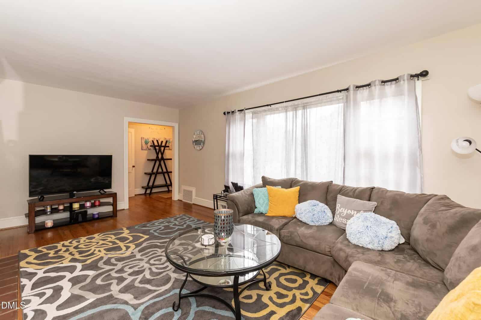 A bright living room with a large gray sectional sofa decorated with yellow and blue pillows, a round glass coffee table, patterned rug, TV on a stand, and sheer curtains on a wide window letting in natural light.