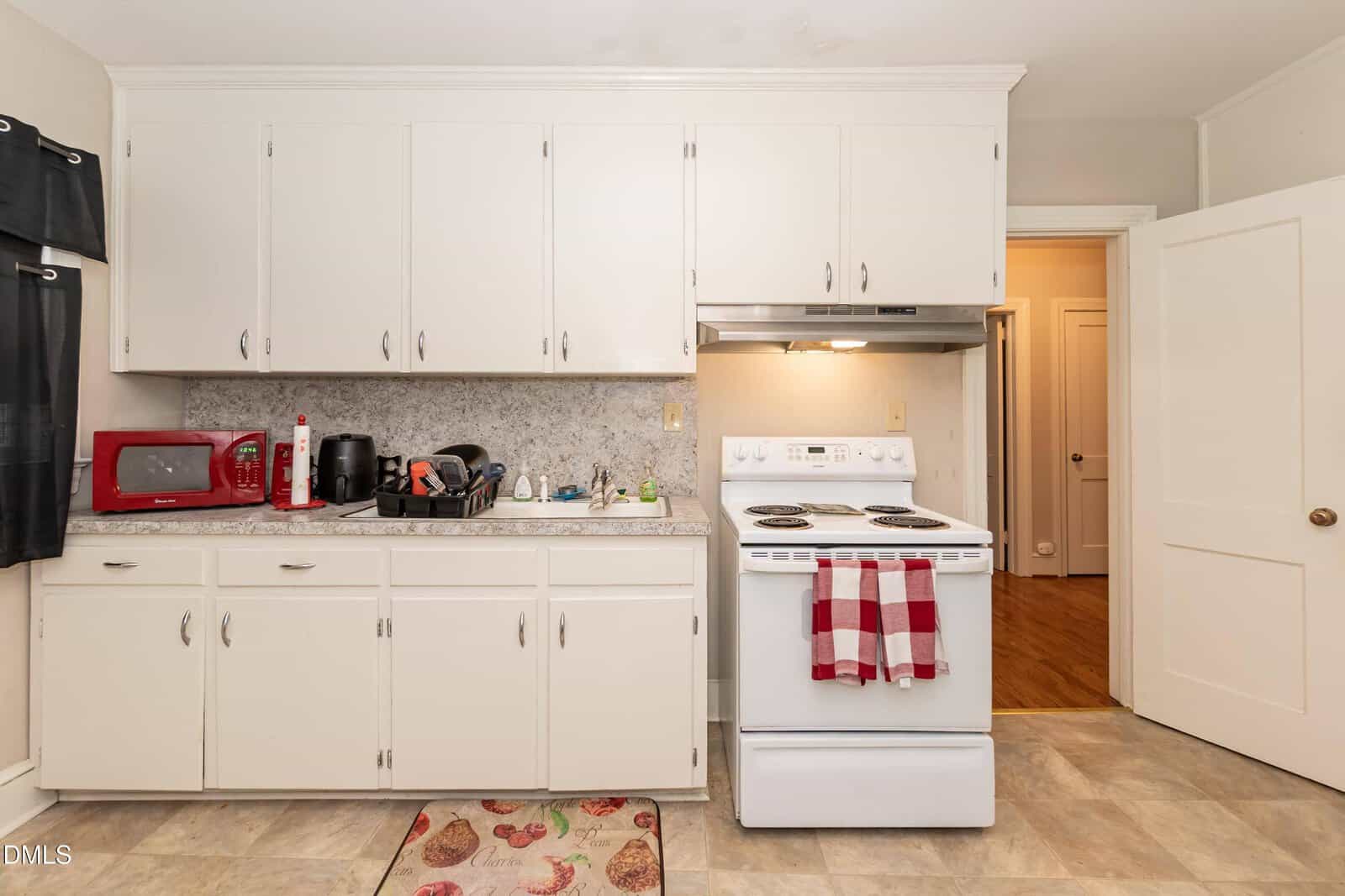 A small kitchen with white cabinets, a white stove with a red plaid towel, a countertop with a drying rack, microwave, and coffee maker, and a mat with fruit near the sink. The floor has beige tiles.