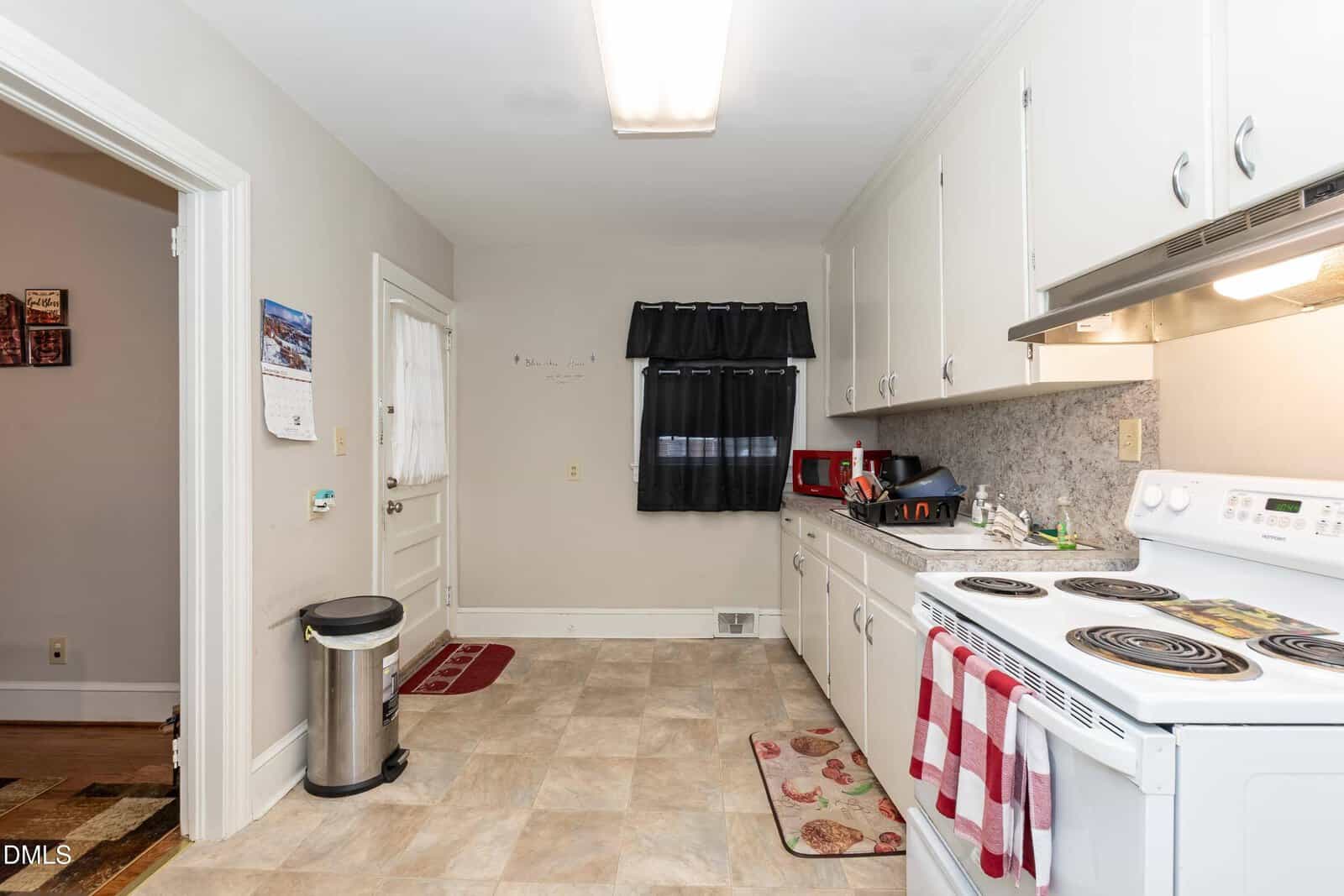 Bright kitchen with white cabinets, white stove, countertop appliances, and a black curtain over a window. There’s a wall calendar, trash can, and two red-striped towels hanging on the oven.
