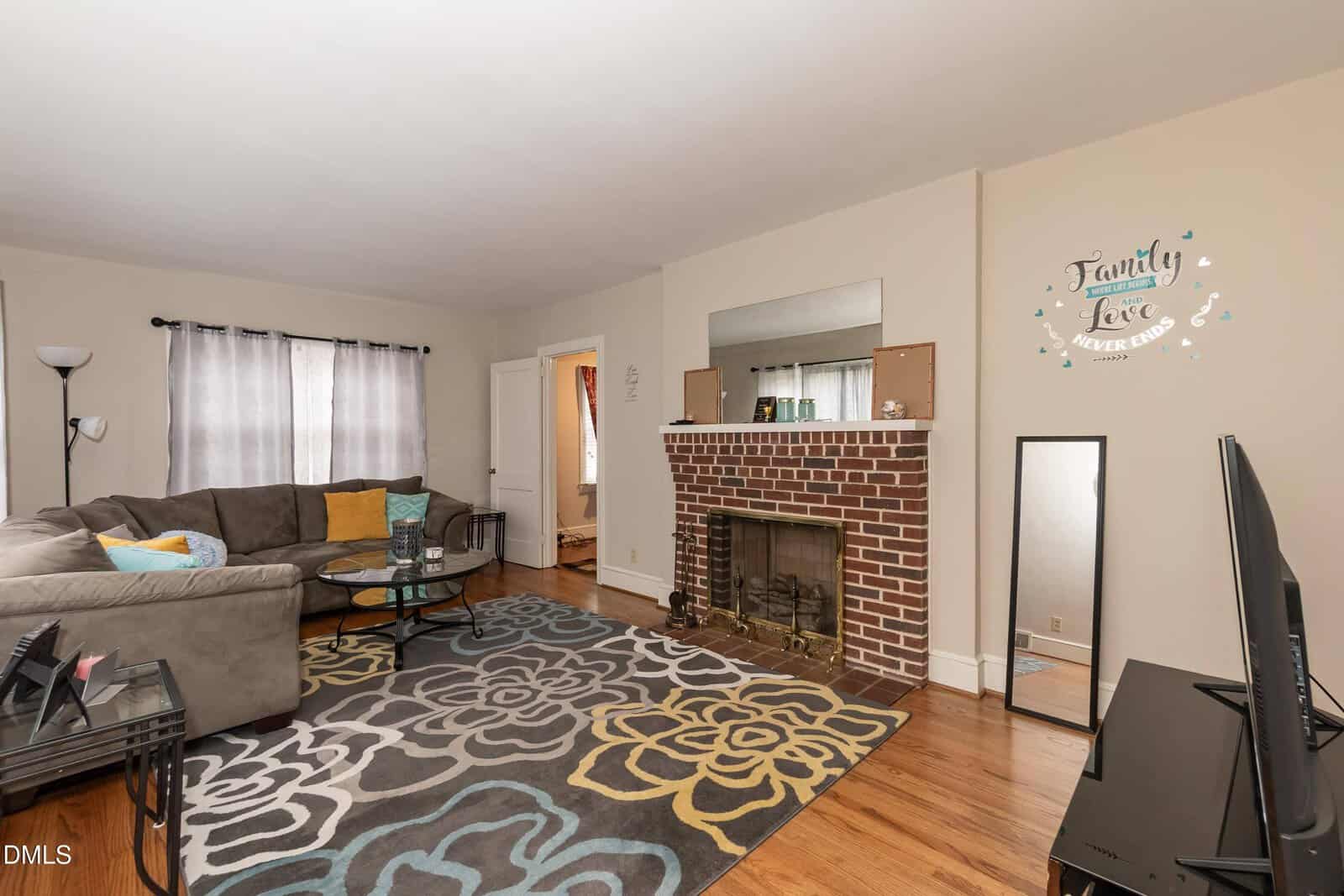 A cozy living room with a sectional sofa, patterned rug, coffee table, TV, brick fireplace, and a wall decal that reads "Family, Love Never Ends." Light-colored walls and wood floors create a warm, inviting space.