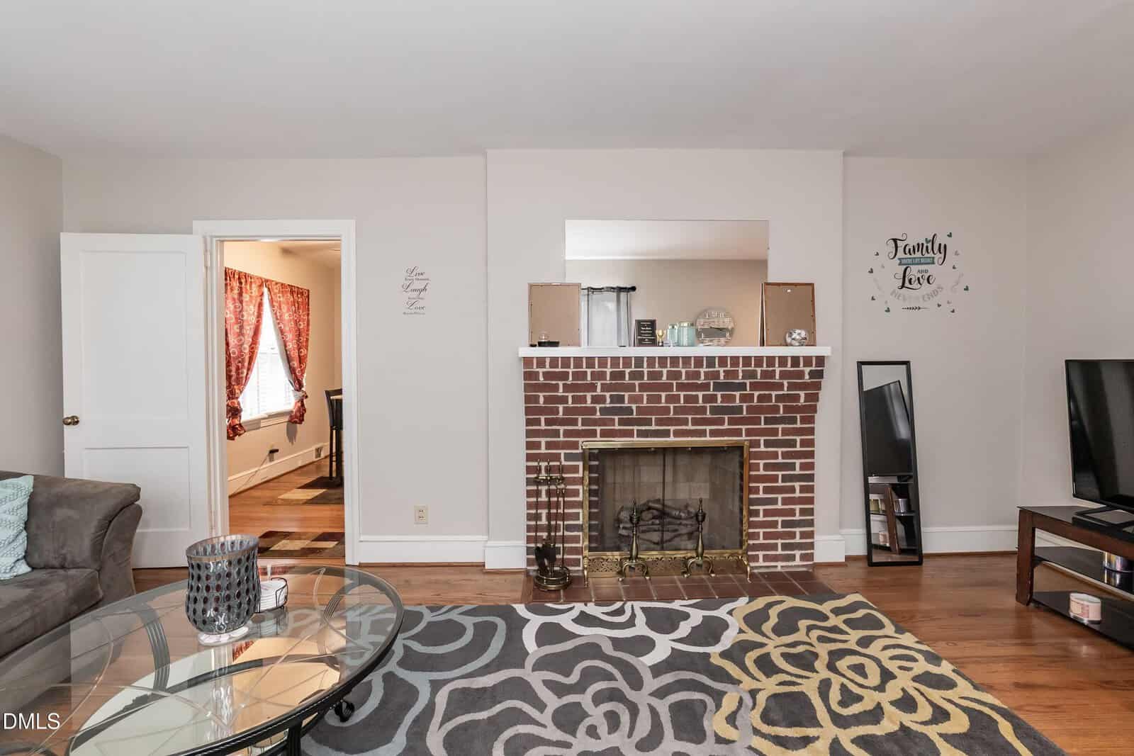 A cozy living room with a brick fireplace, a glass coffee table, a patterned rug, and a TV. A doorway leads to another room with red curtains. Wall art and decor sit above the fireplace. Hardwood floors throughout.