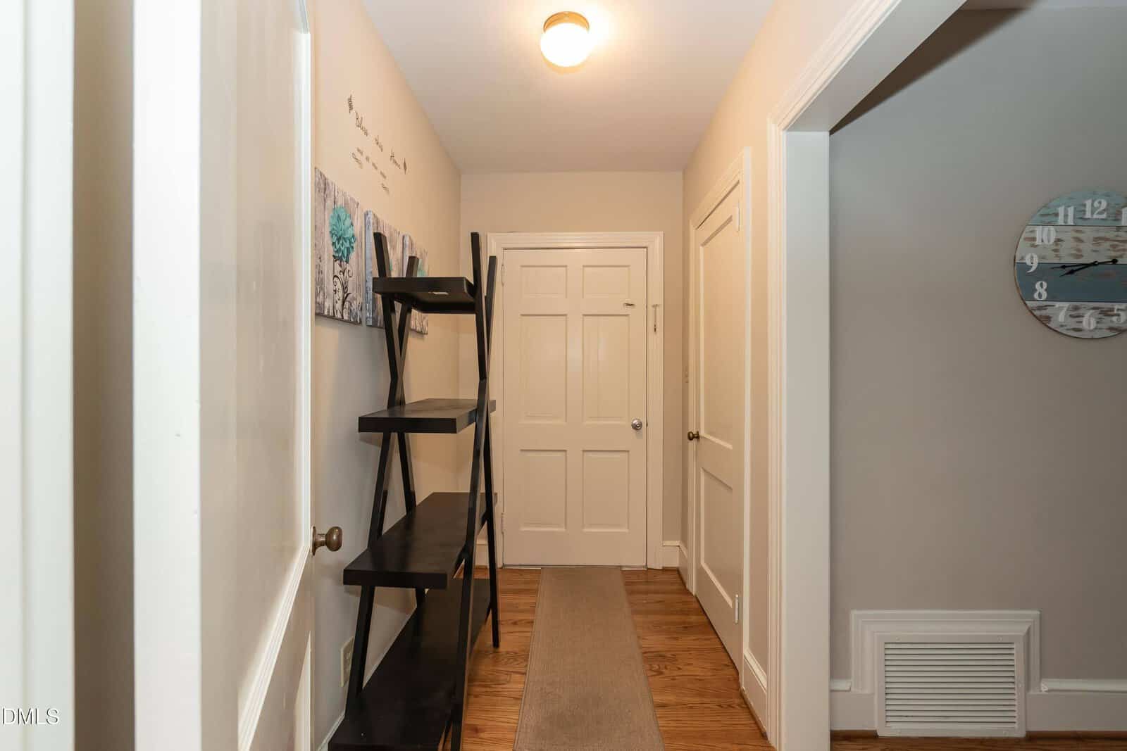A hallway with light-colored walls, wooden floor, a black ladder-style shelf on the left, two white doors at the end, a wall clock on the right, and a ceiling light fixture.