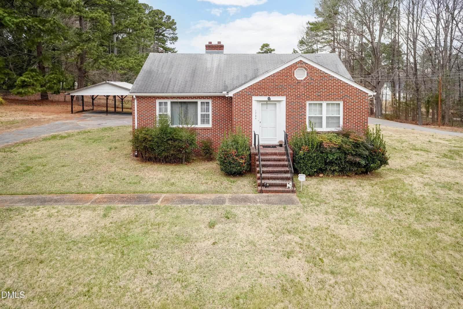 A single-story red brick house with a white door, front steps, and bushes lining the front. The house sits on a grassy lawn with a driveway leading to a detached double carport. Trees surround the property.