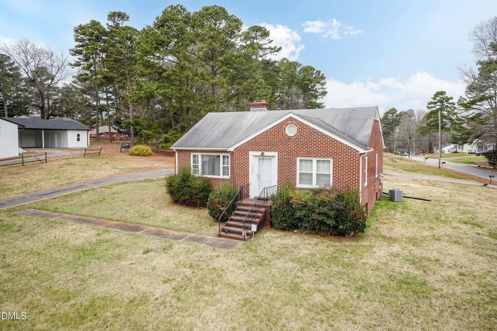 A small red-brick house with white trim and a gray roof sits on a grassy lot with bushes by the front steps, surrounded by trees and neighboring homes under a partly cloudy sky.