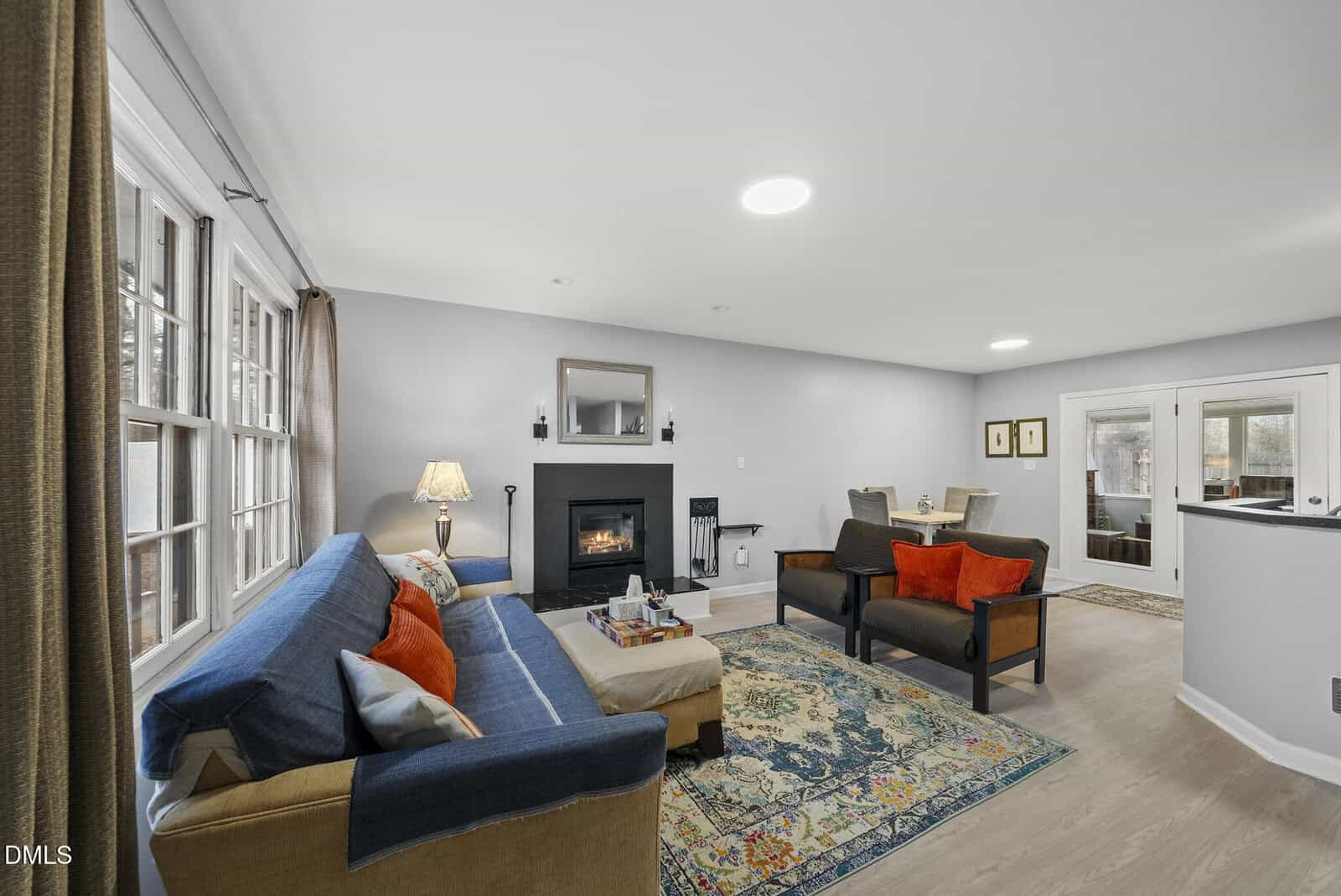 A bright, cozy living room with a blue sofa, armchairs, colorful cushions, a fireplace with a mirror above, a patterned rug, and large windows letting in natural light. Dining area visible in the background.