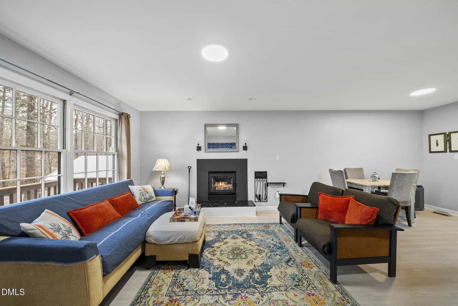 A bright living room with large windows, a blue sofa, two armchairs with orange cushions, a fireplace, a patterned rug, and a dining table with four chairs in the background.