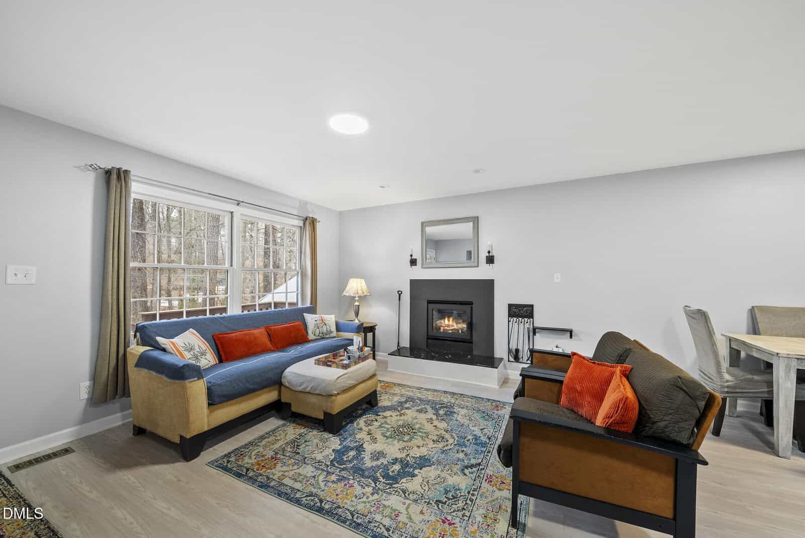 A cozy living room with a blue sectional sofa, orange and brown cushions, a patterned rug, a fireplace with a mirror above it, a wooden chair, and a dining table in the background. Large windows let in natural light.