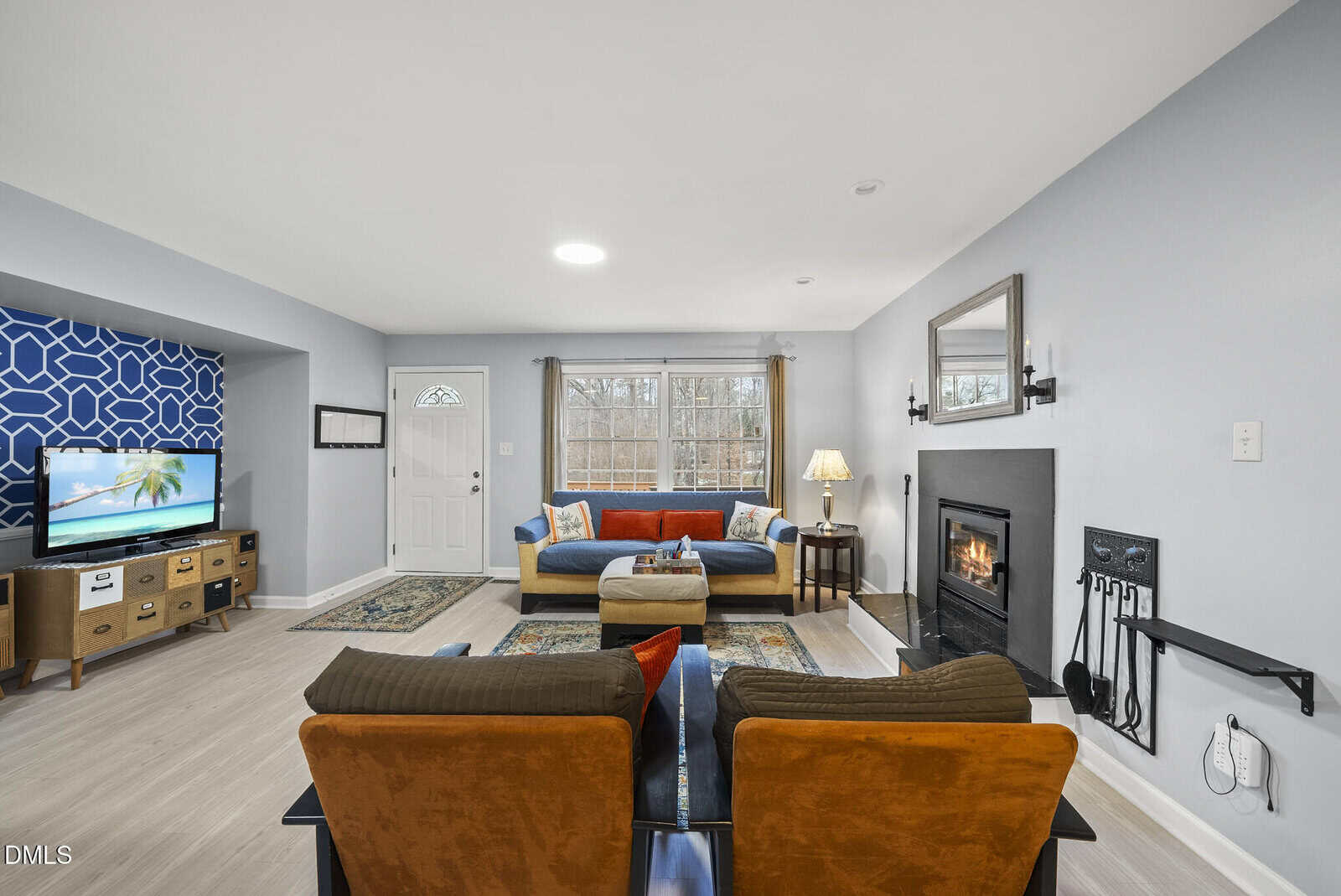 A bright living room with a sofa, two armchairs, a TV on a wooden stand, a fireplace with tools, a wall mirror, area rugs, and large window. Light-colored walls and flooring create an airy feel.
