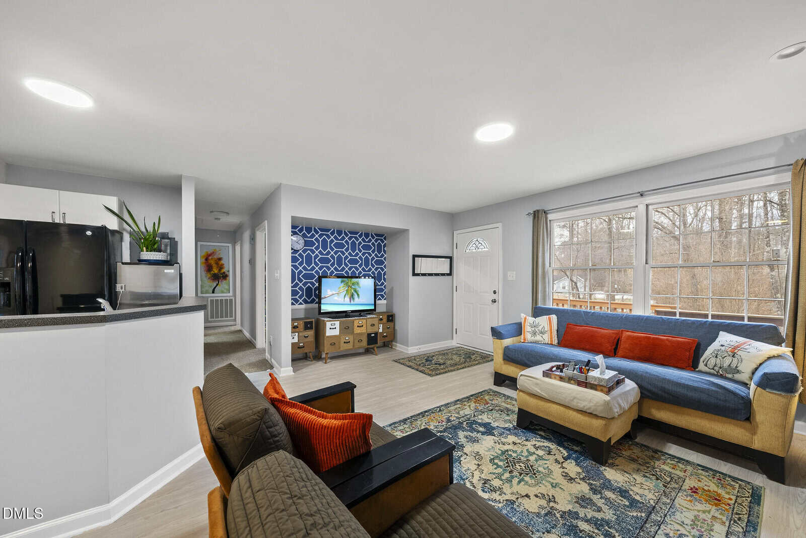 A bright, modern living room with large windows, a sectional sofa with red and blue cushions, a TV on a console with a geometric blue accent wall, and an open kitchen with white cabinets and black appliances.