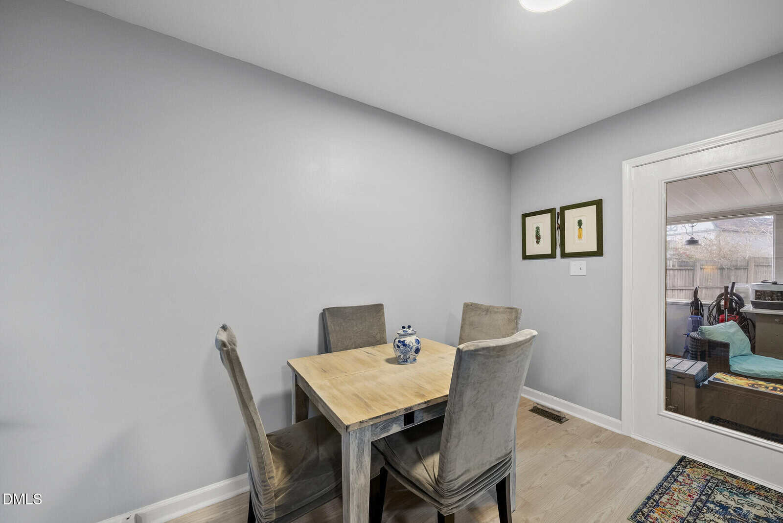 A small dining area with a light wood table and four gray upholstered chairs, a blue and white vase on the table, and two framed prints on a pale gray wall near a glass door leading outside.