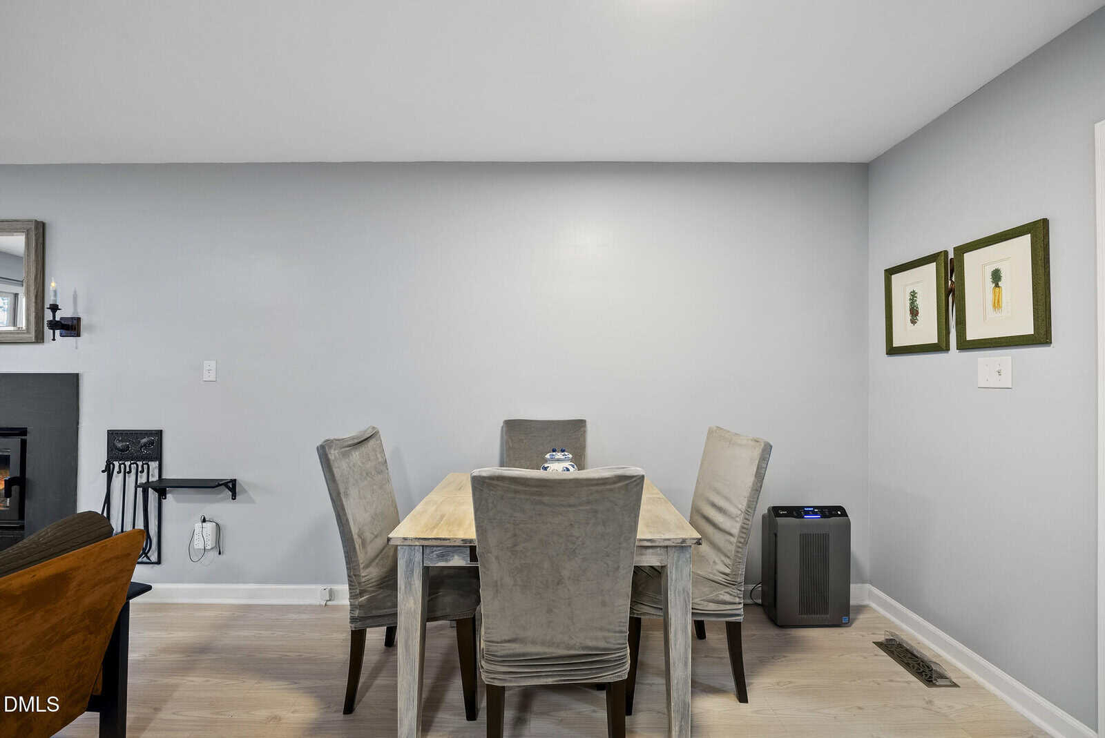 A dining area with a light wood table and four gray upholstered chairs sits against a light gray wall. Two framed artworks hang on the right wall. An air purifier is on the floor nearby.