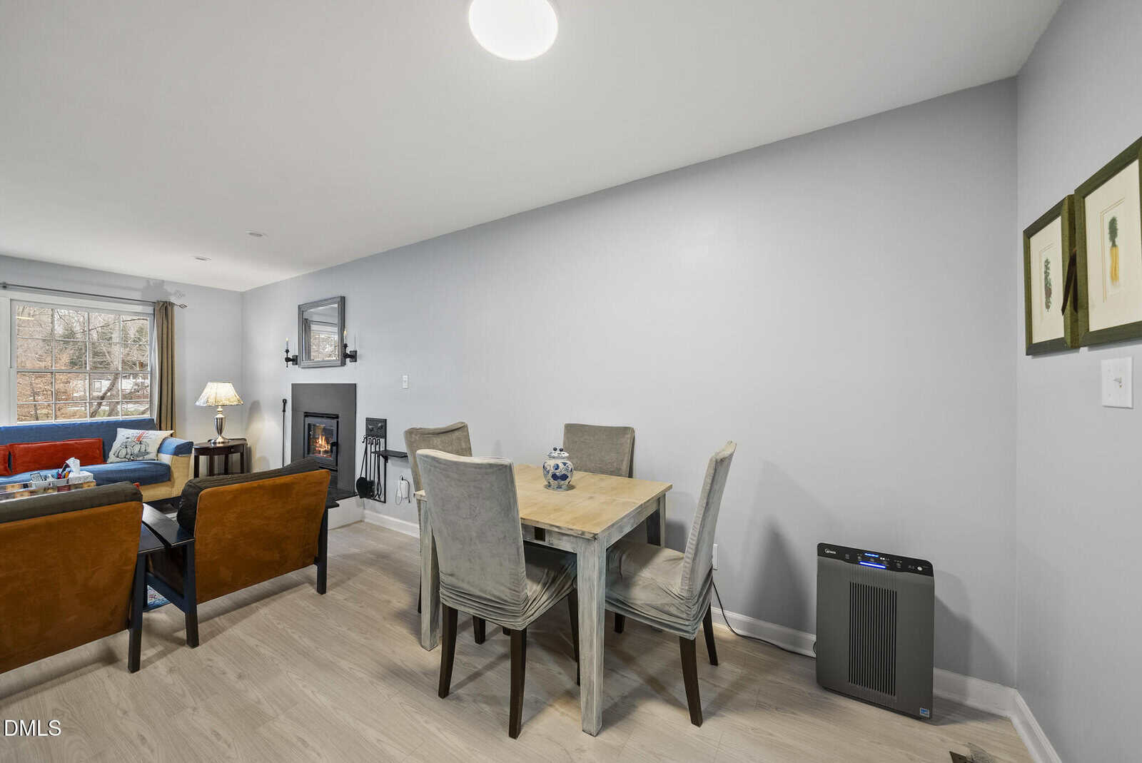 A modern living and dining area with light gray walls, wood flooring, a small dining table with four chairs, two armchairs, a sofa, a fireplace, framed art, and a window letting in natural light.