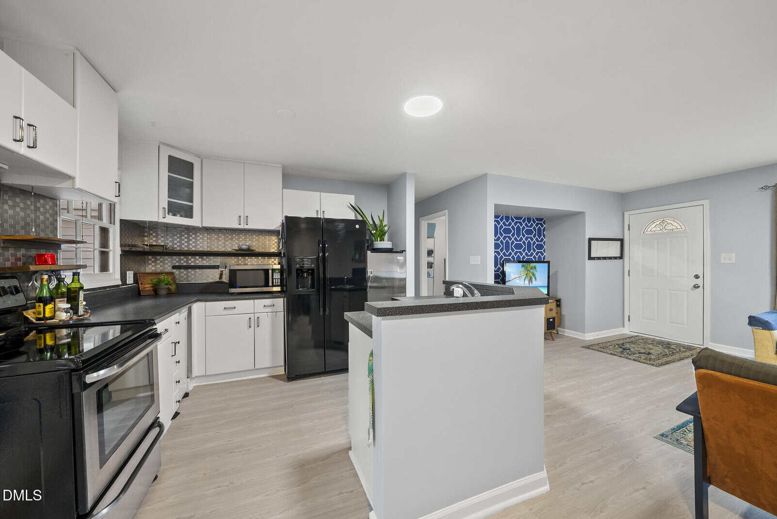 Modern kitchen with white cabinets, black appliances, and light wood flooring, open to a living room with a patterned accent wall, TV, and front door, all brightly lit with natural and ceiling light.