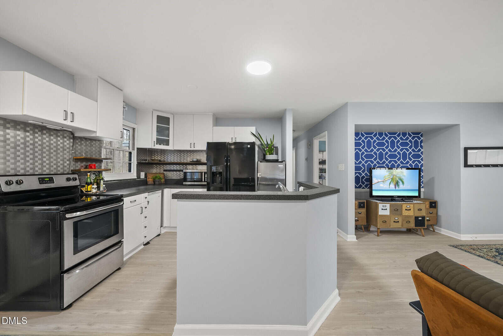Modern kitchen with white cabinets, stainless steel stove, black appliances, and patterned backsplash. Open layout leads to a living area with a TV on a wooden stand and blue geometric accent wall. Light wood flooring throughout.